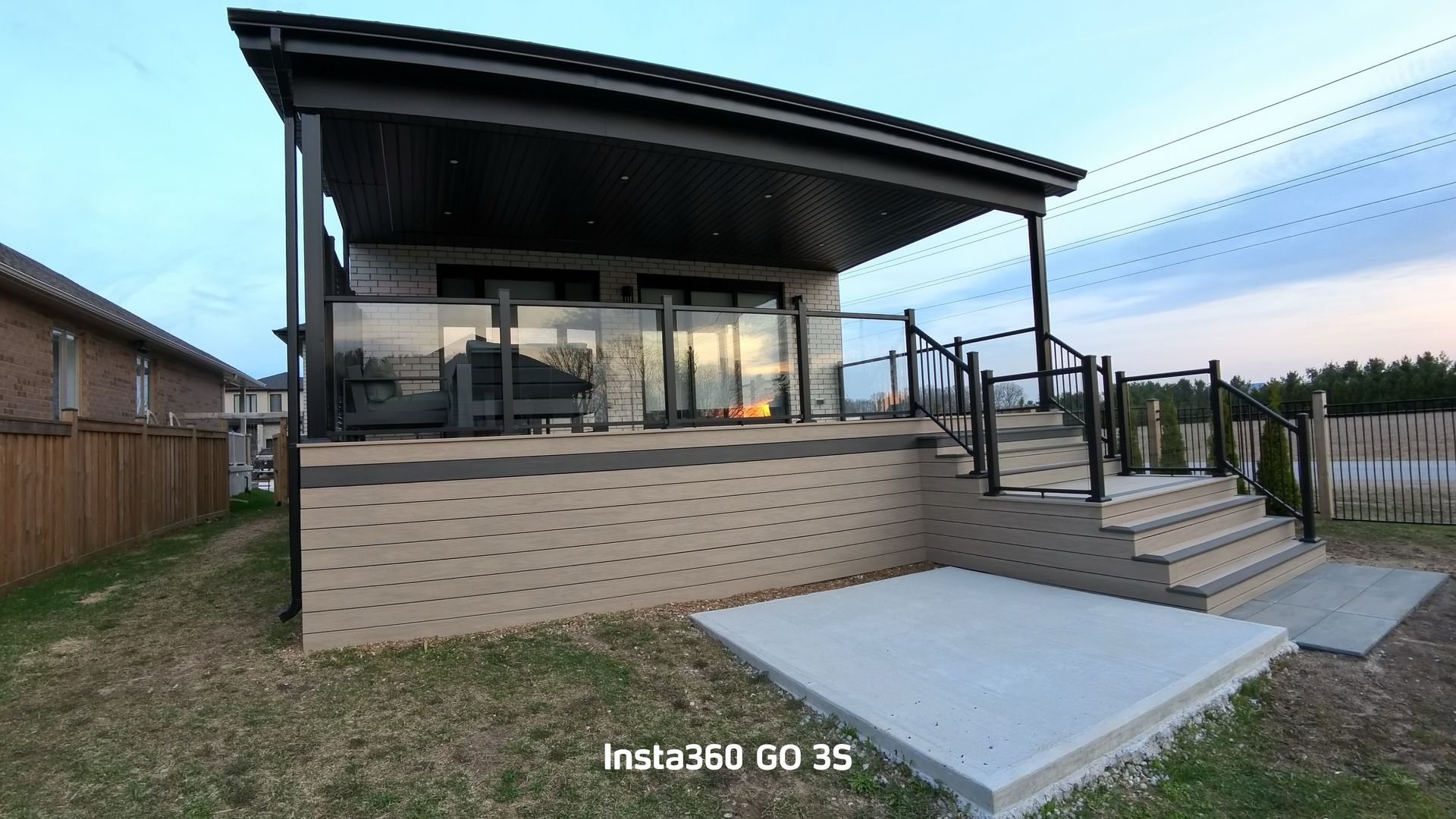 Backyard patio with a covered deck, stairs, glass railing, and concrete patio. Cloudy sky.