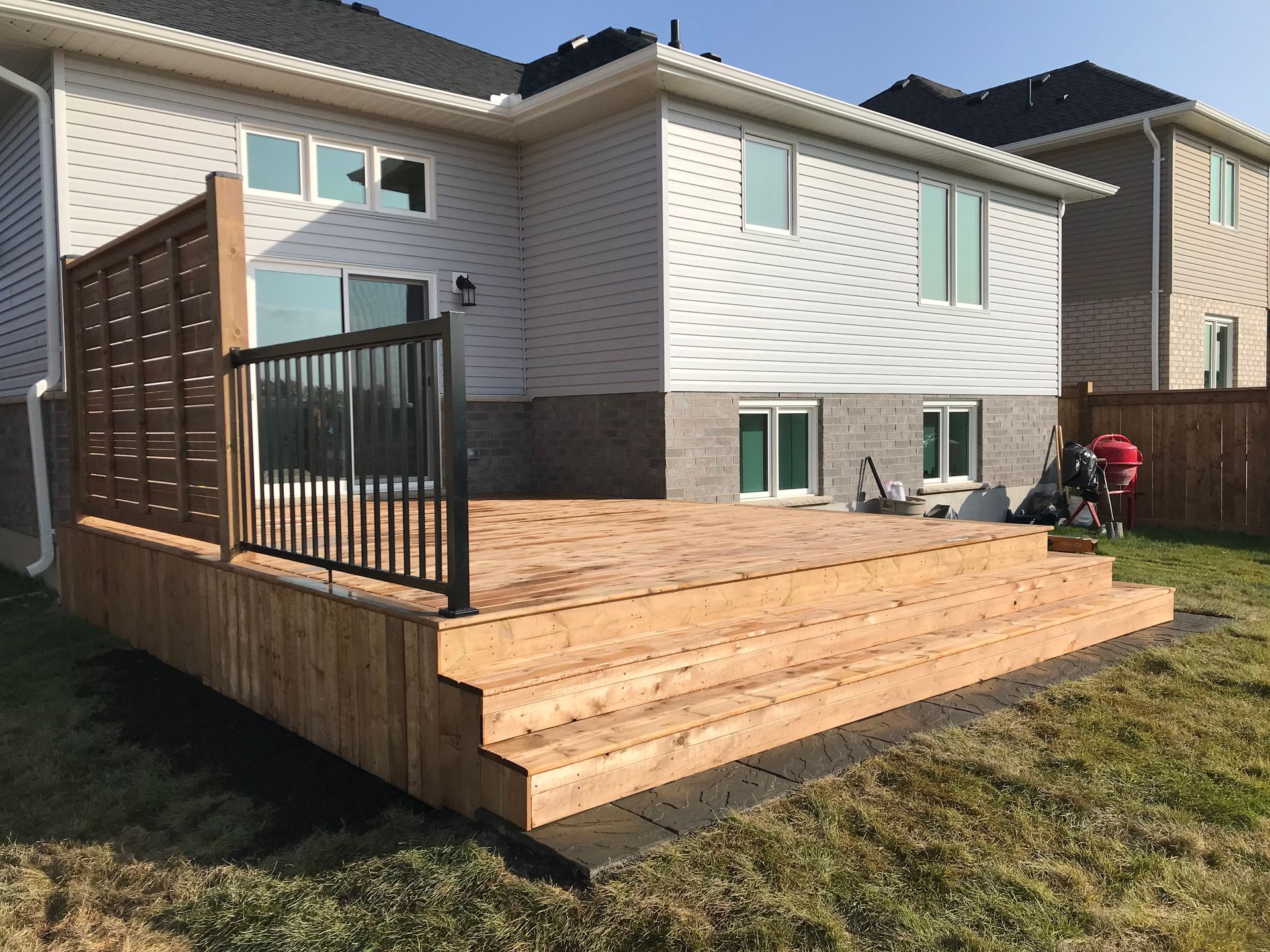 Wooden deck with steps, a black metal railing, and a privacy screen attached to the side of a house.