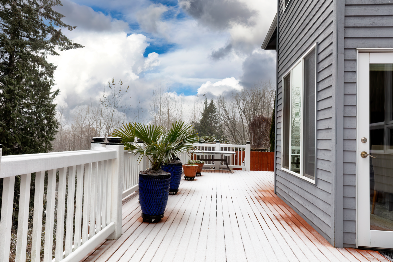 Snow-covered wooden deck with white railing and potted plants, connected to a gray house, overcast sky.
