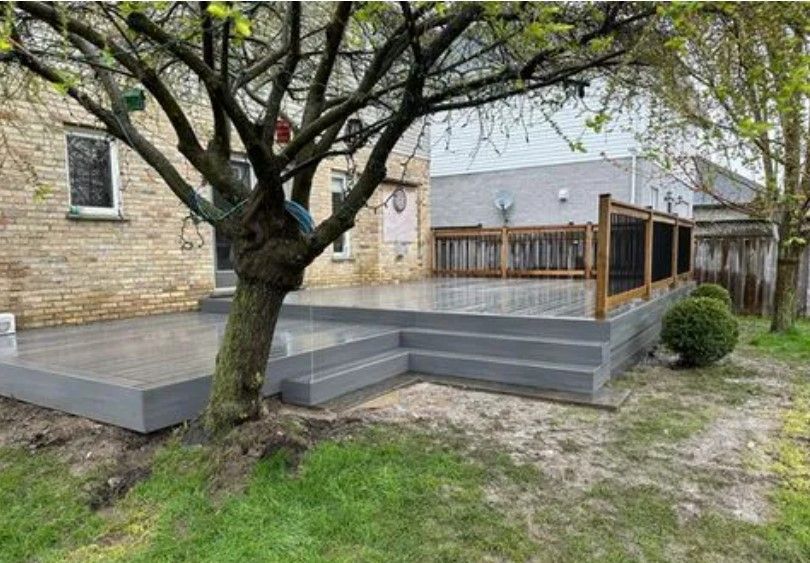 Concrete patio with steps, bordered by a wooden railing and a small tree. Grey tones, overcast day.