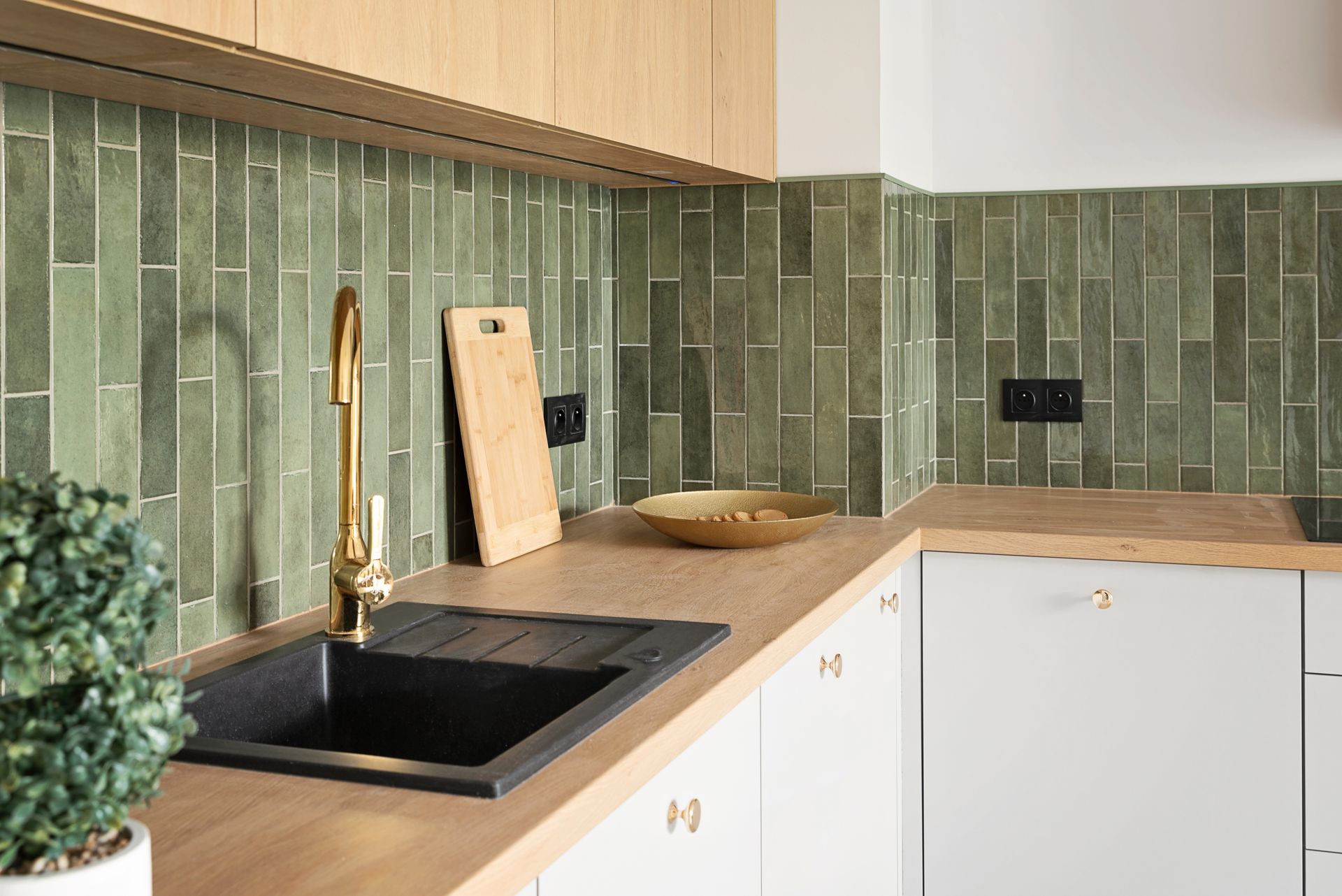 Modern kitchen corner with black sink, brass faucet, green tile backsplash, and light wood counters