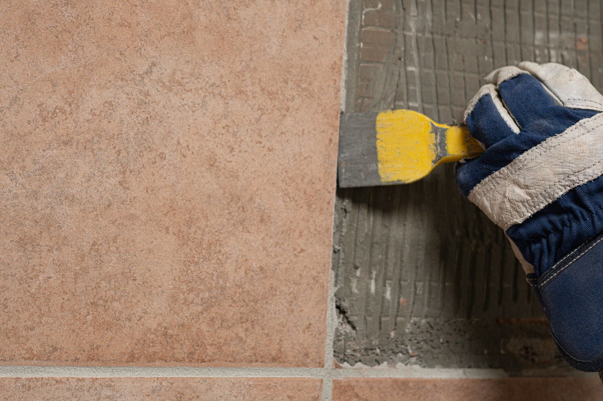 Gloved hand using a yellow trowel to spread adhesive on floor tile surface