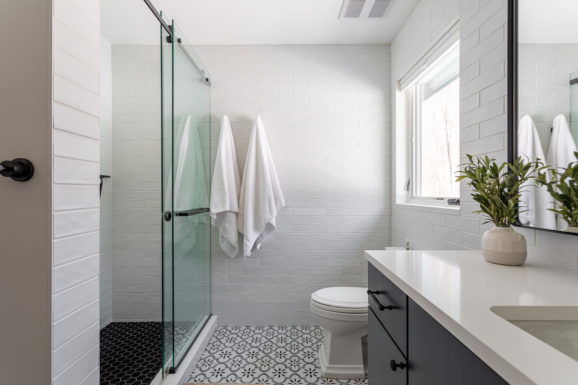 Modern white bathroom with glass shower, patterned floor, vanity, towels, and window