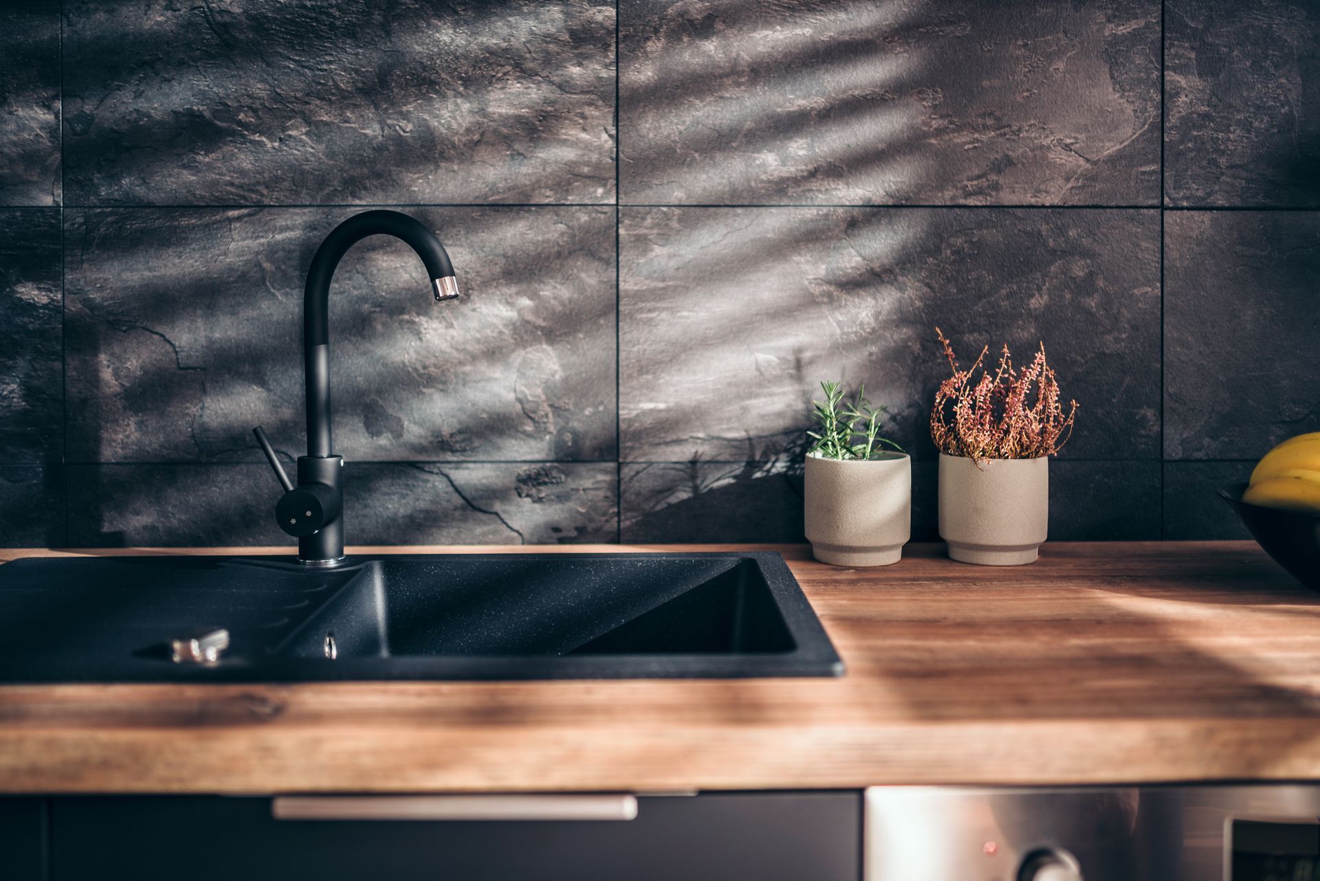 Modern kitchen counter with black sink, wooden countertop, potted plants, and striped sunlight on tiled backsplash