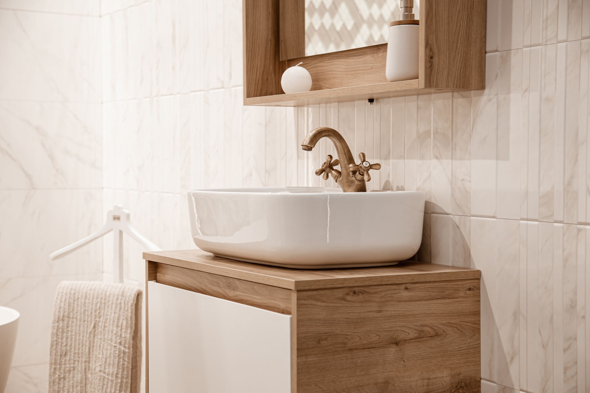 Wooden bathroom vanity with white vessel sink, brass faucet, and a mirrored shelf above, in a bright tiled room