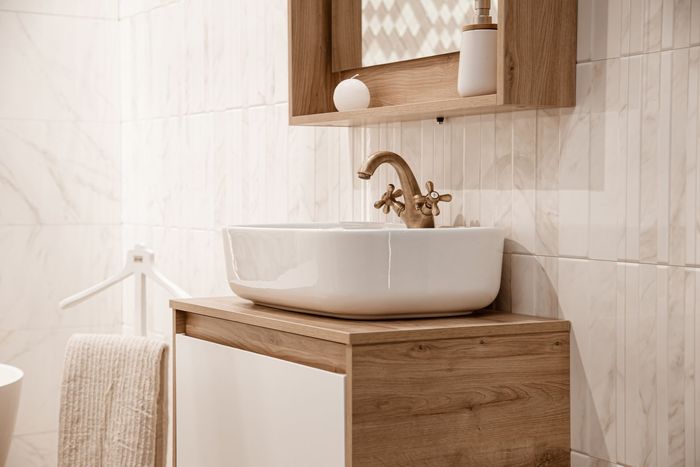 Wooden bathroom vanity with white vessel sink, brass faucet, and a mirrored shelf above, in a bright tiled room