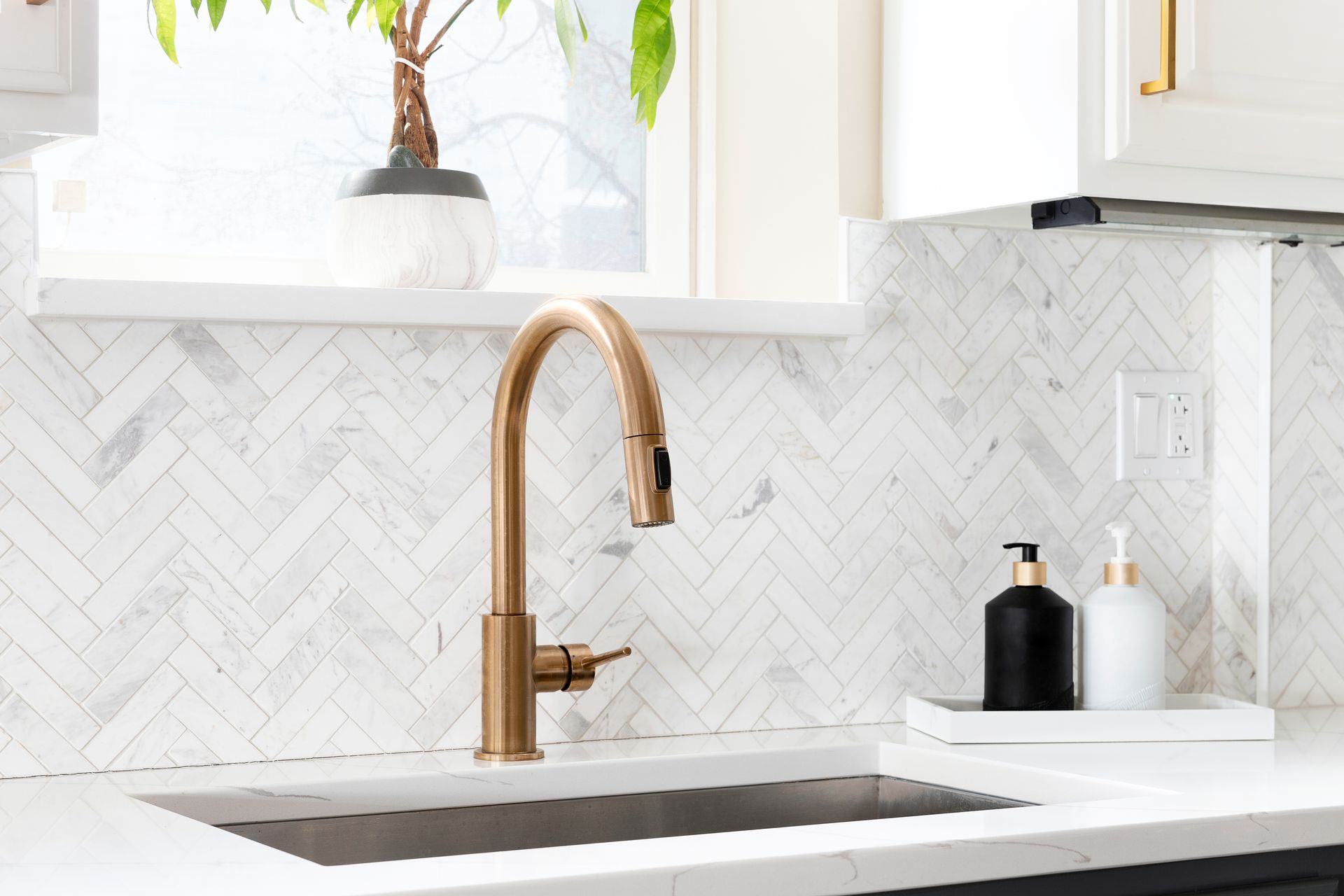 Modern kitchen sink with brass faucet, white herringbone backsplash, and soap dispensers on a tray