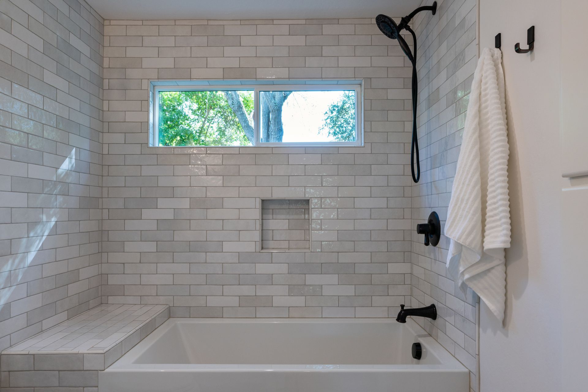 Modern white-tiled shower with tub, window, black fixtures, and towel on a wall hook.