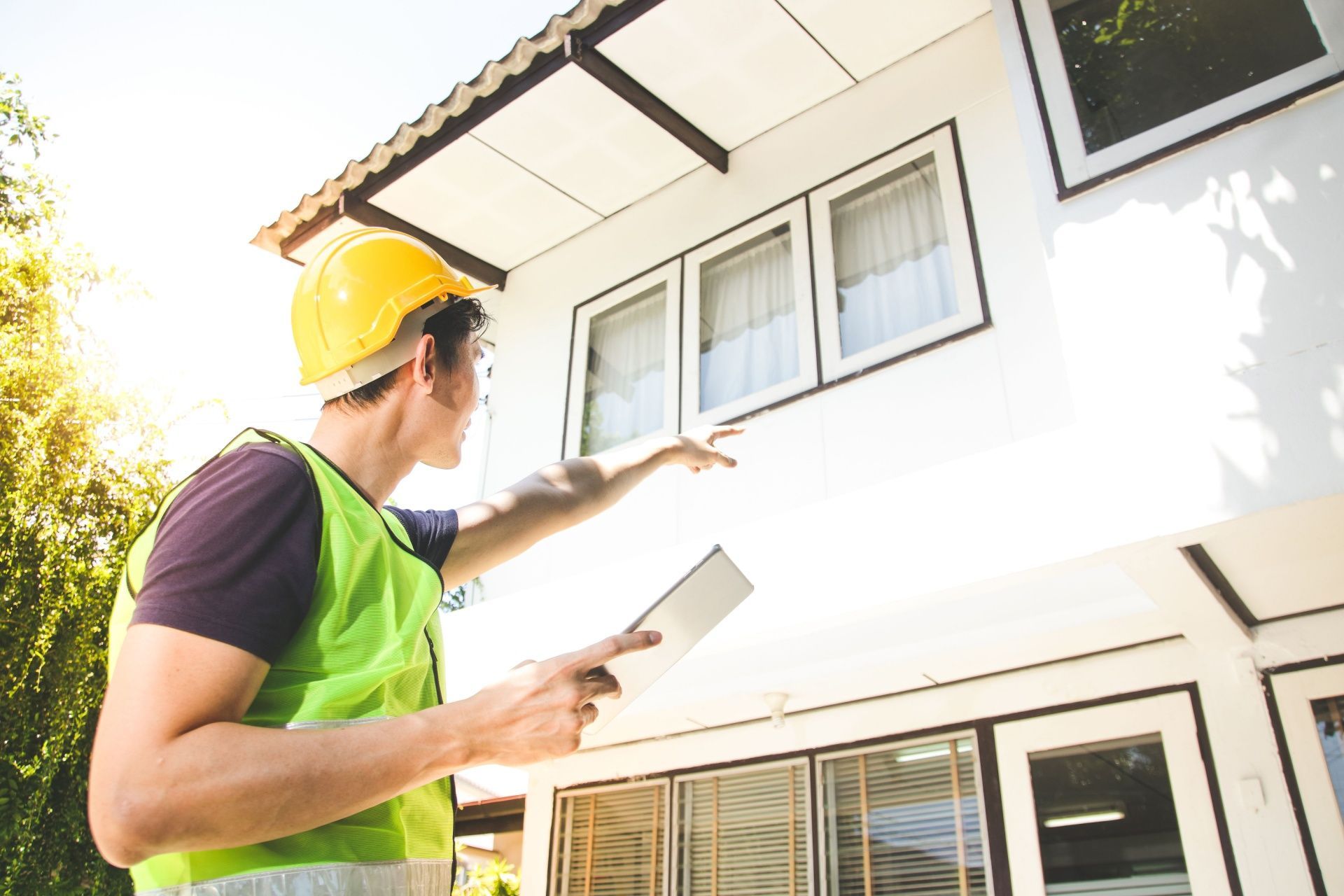 Worker in a yellow hard hat and green vest pointing at a house exterior while holding a tablet