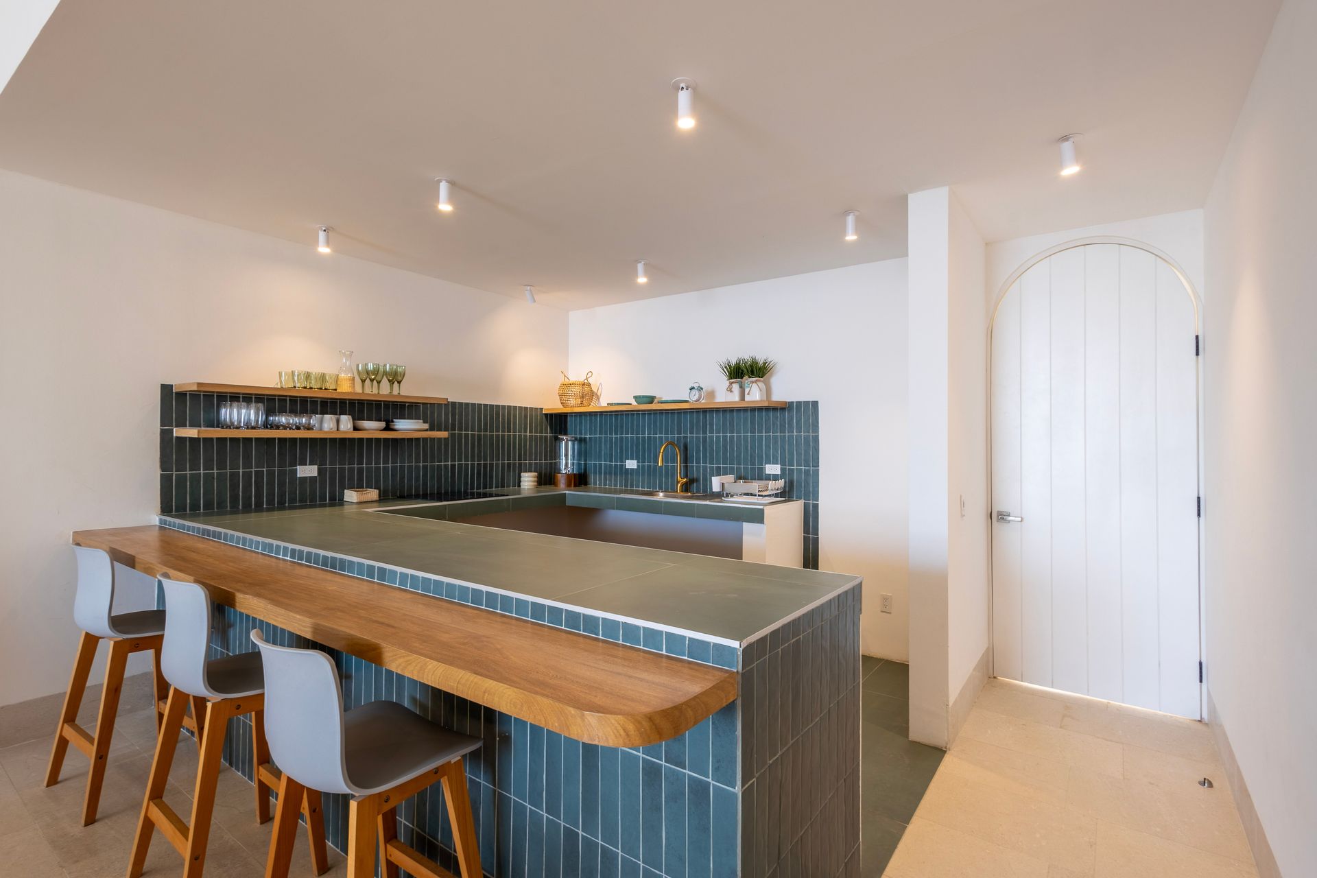 Modern kitchen with wood bar, blue tiles, and three gray stools. White walls and a door are visible.