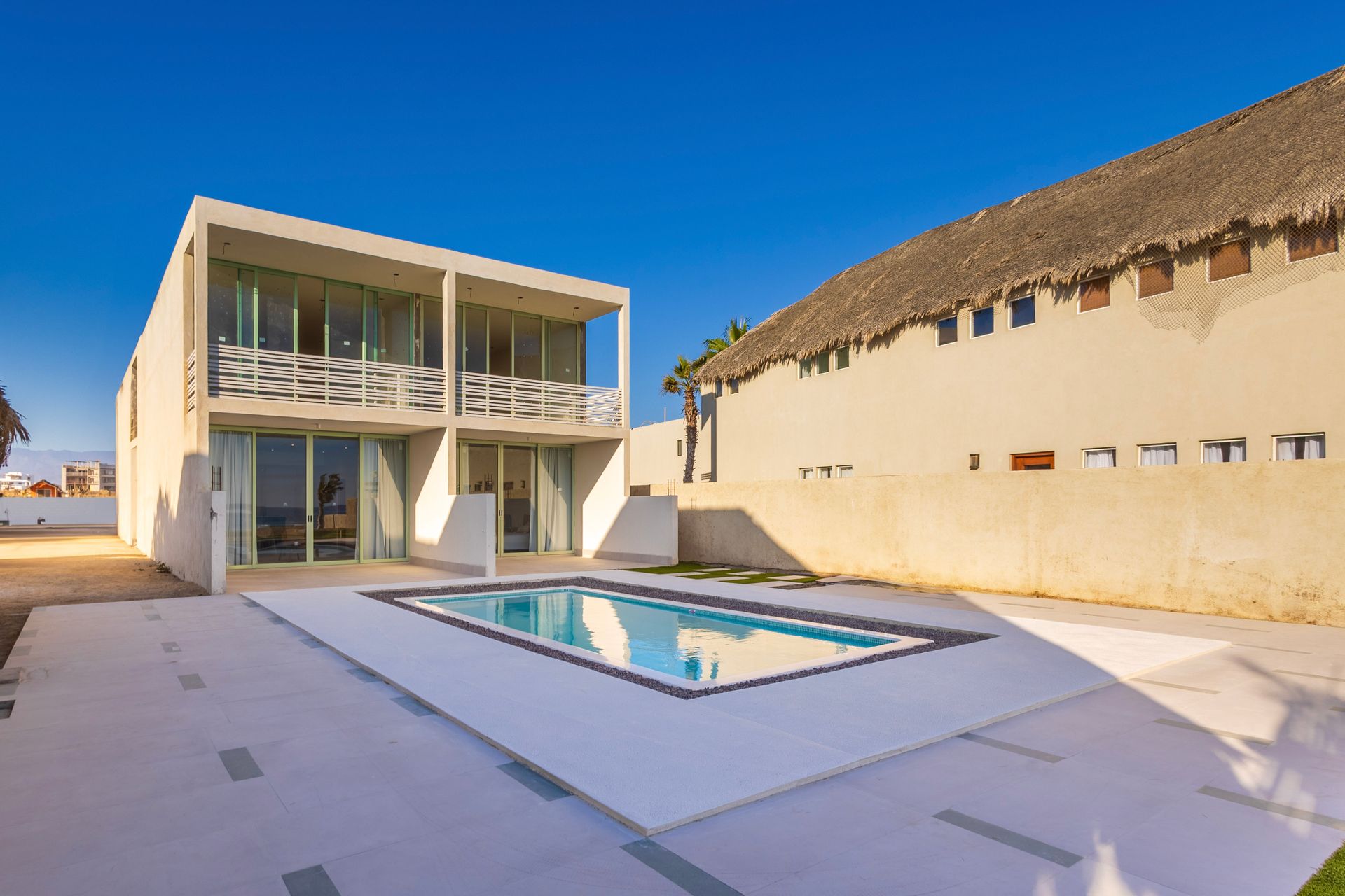 Modern two-story house with pool, next to a building with a thatched roof, on a sunny day.