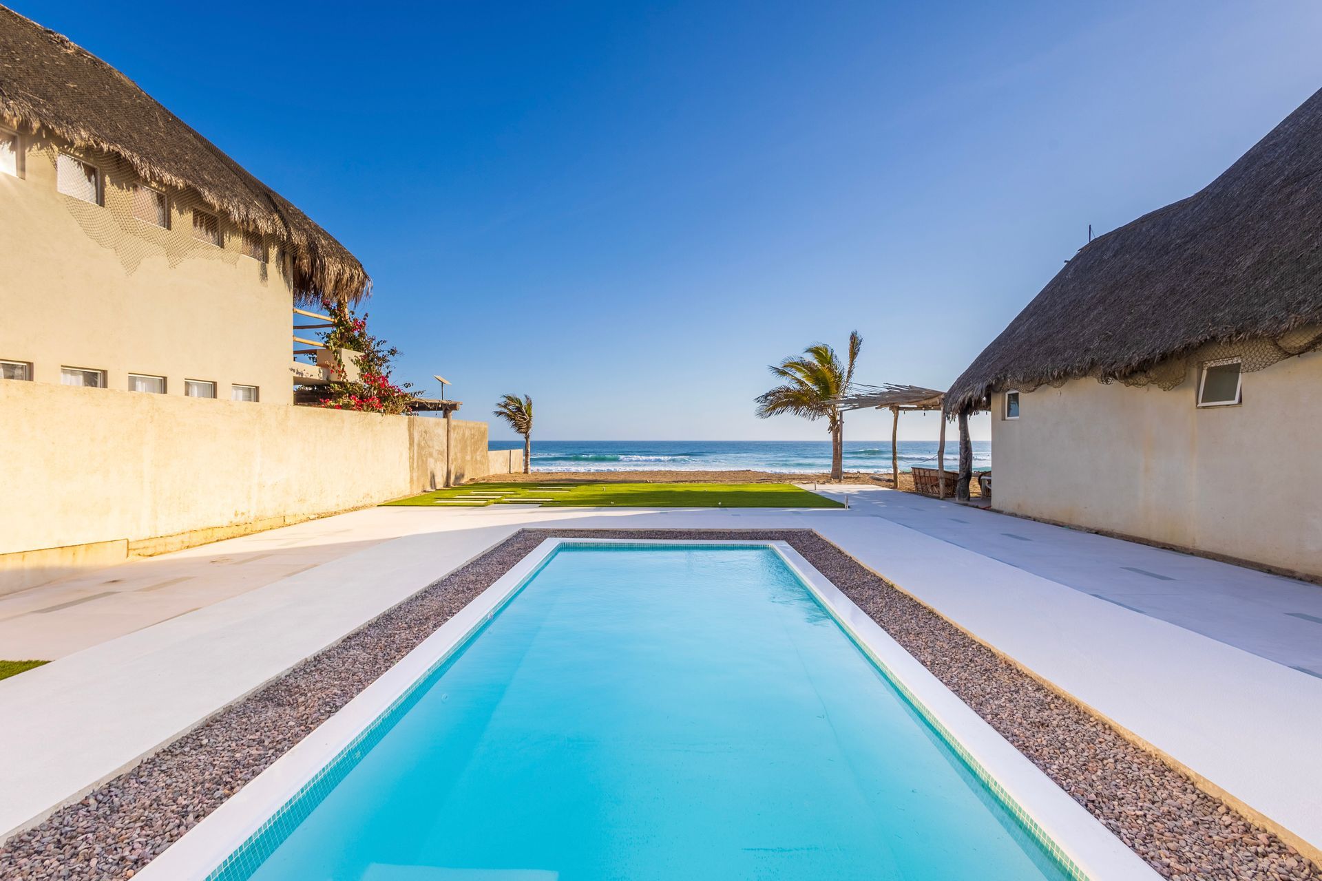 Pool between two buildings with thatched roofs, overlooking the ocean under a blue sky.