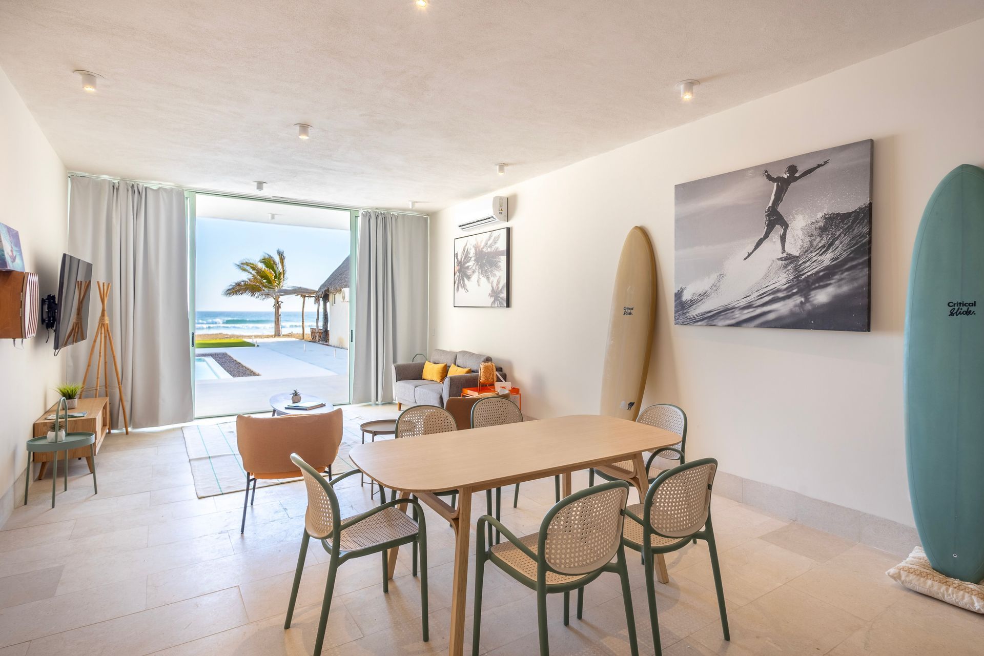 Bright, open living area with ocean view. Features a dining table, surfboards, and a surfer photo.