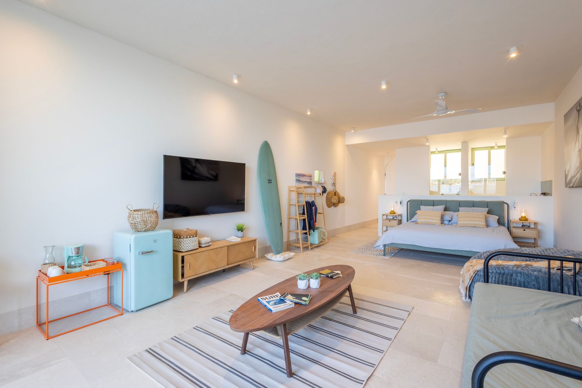 Open-concept living space with a TV, surfboard, bed, and seating area. Light blue and wood tones.