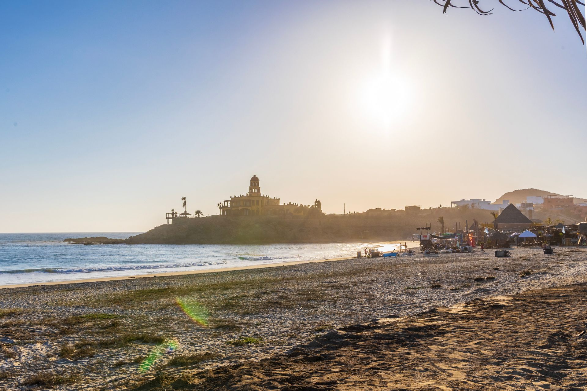 Beach scene at sunset with a stone structure on a small cliff, boats, people, and a bright sun.