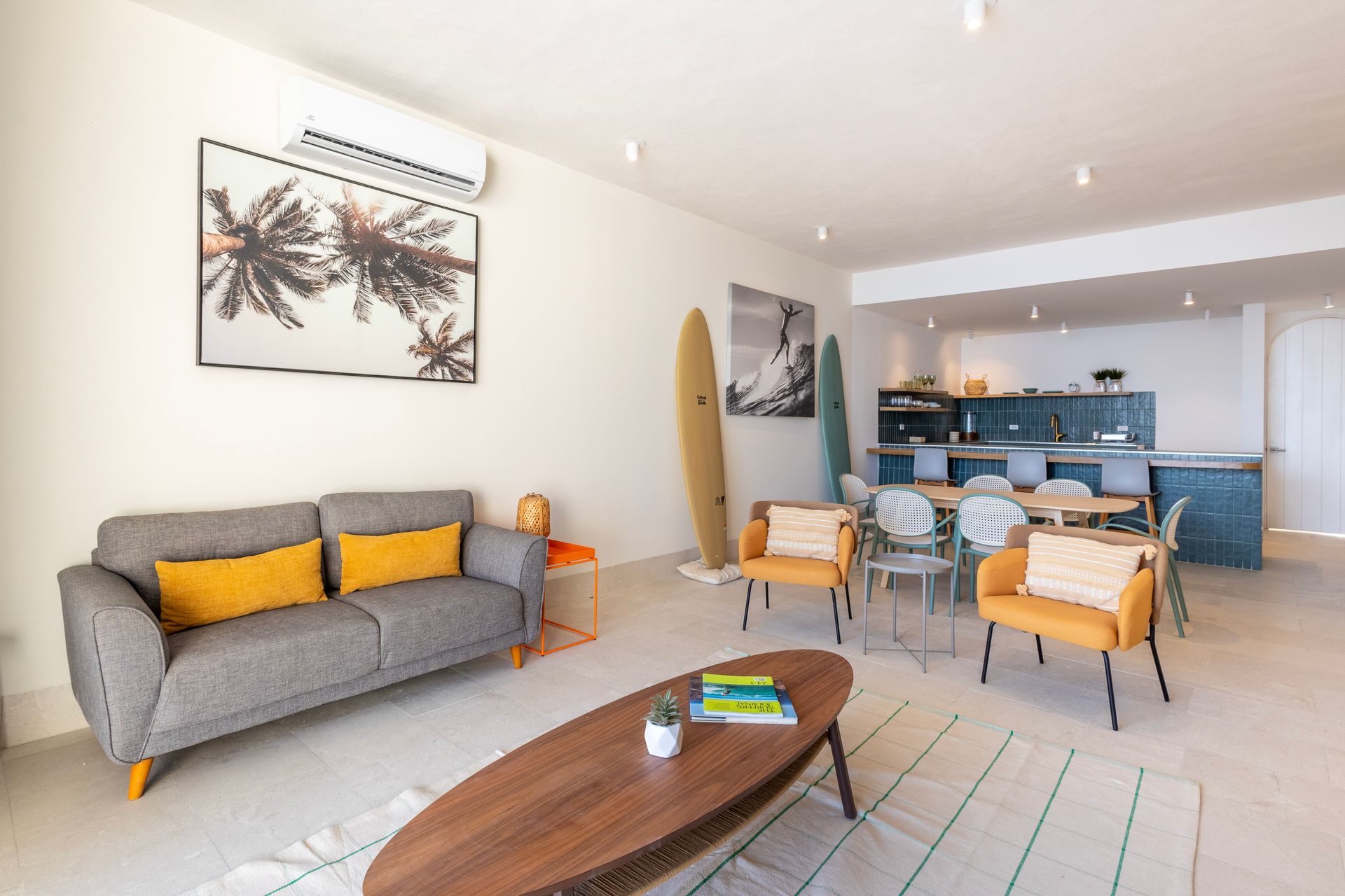 Living room with gray sofa, yellow pillows, surfboards, and orange chairs, in a bright coastal setting.