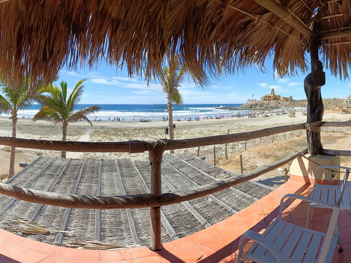 View from a thatch-roofed balcony overlooking a beach with people, ocean, and palm trees under a blue sky.