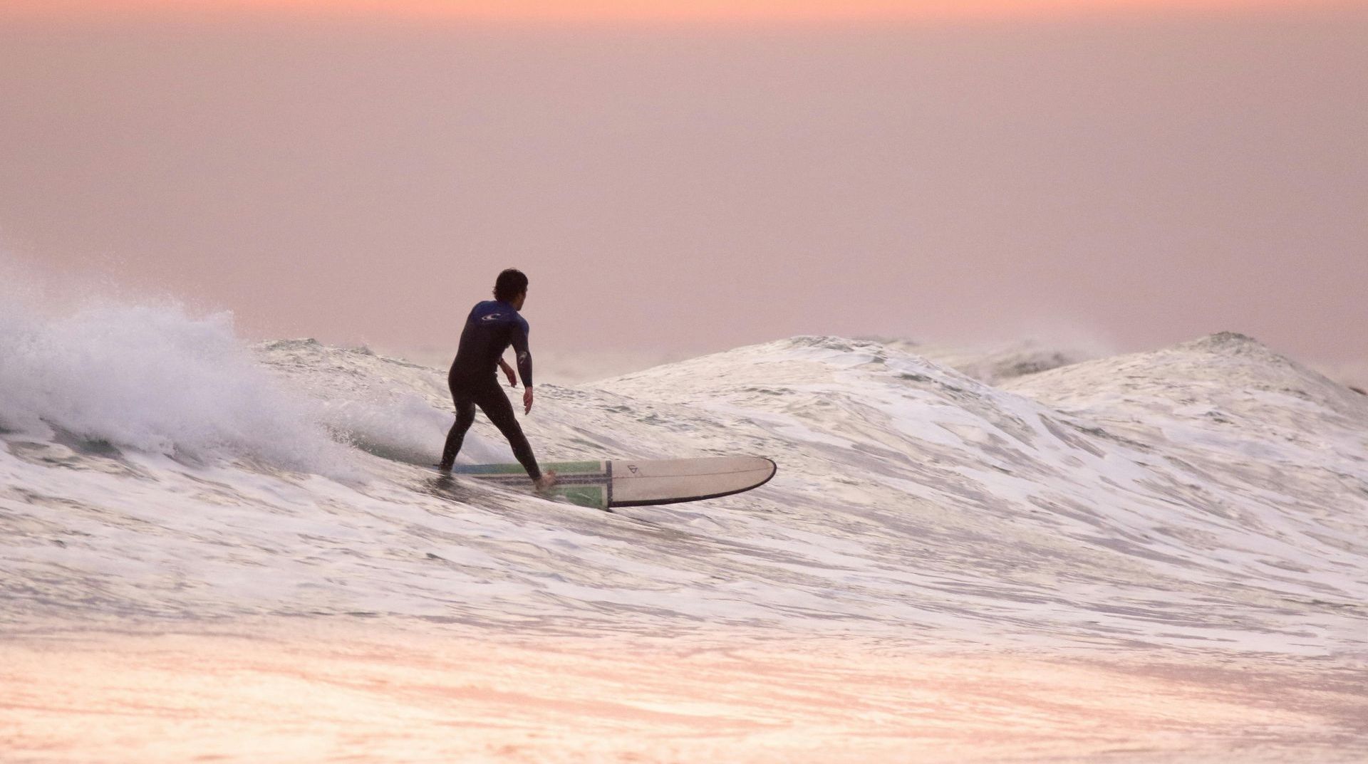 View from a thatch-roofed balcony overlooking a beach with people, ocean, and palm trees under a blue sky.