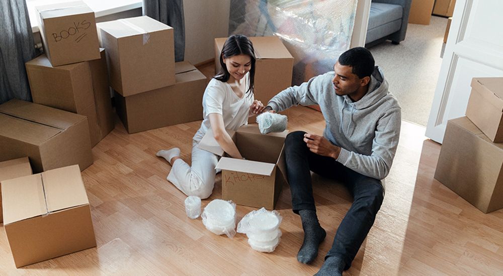 A man and a woman are sitting on the floor in a living room surrounded by cardboard boxes.