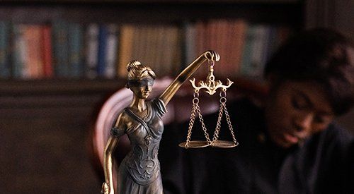 A woman is sitting in front of a statue of lady justice holding scales of justice.