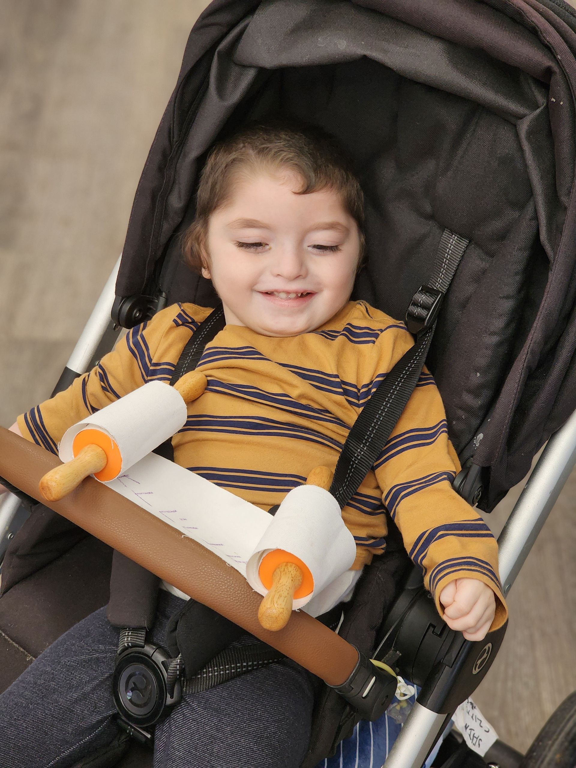 A little girl is sitting in a stroller with a rolling pin attached to the handle.