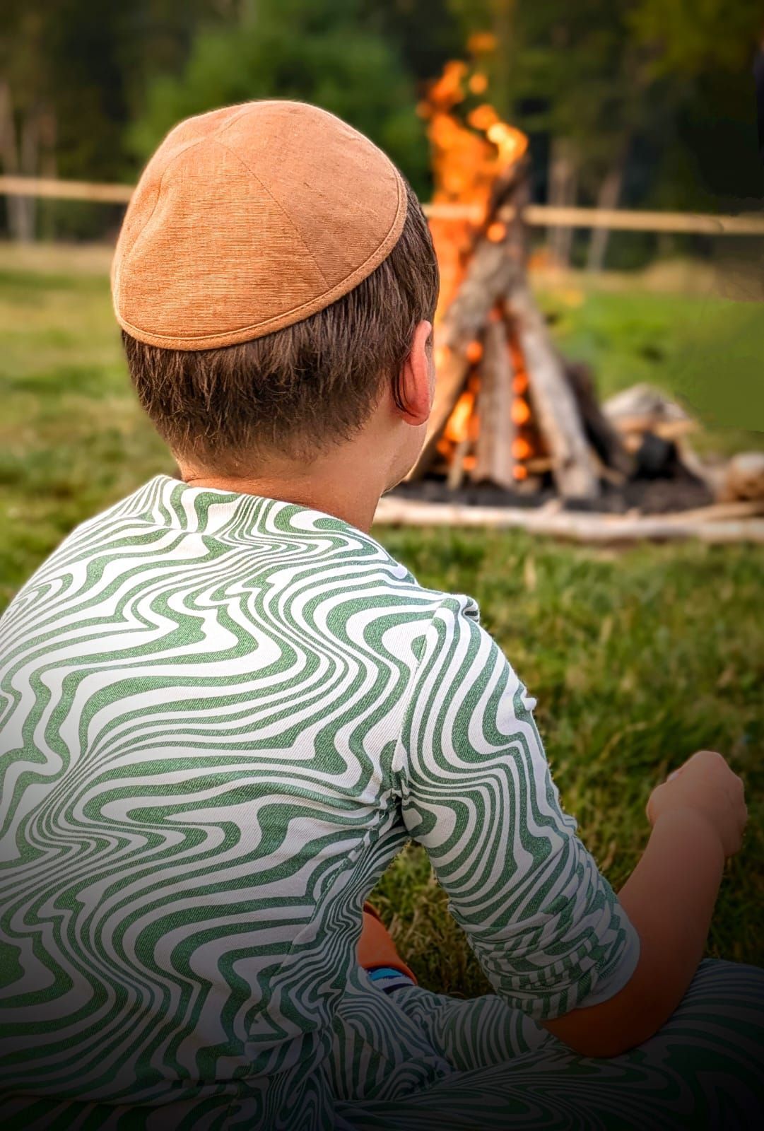 A young boy wearing a jewish hat is sitting in front of a campfire.
