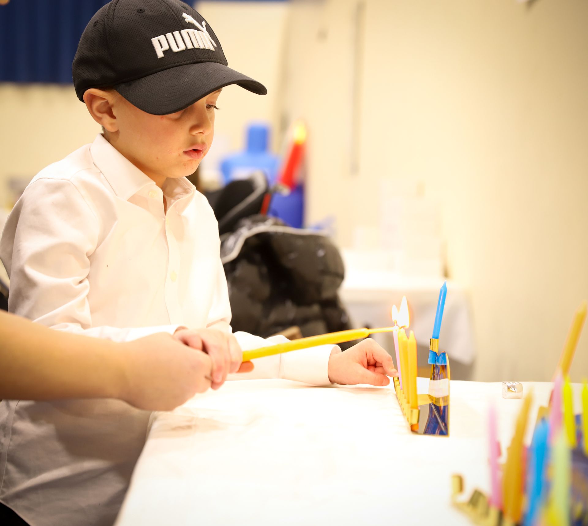 A young boy wearing a puma hat is lighting a menorah.