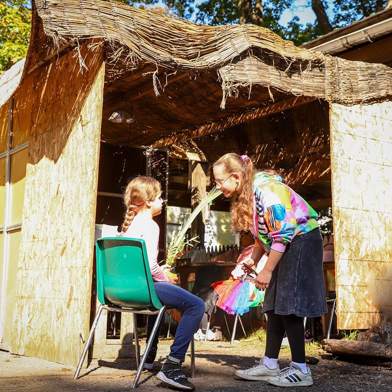 A girl in a tie dye shirt is standing next to a young girl in front of a sukkah.
