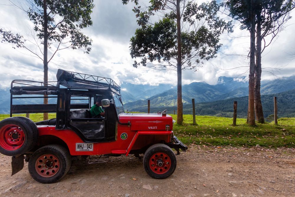 Jeep rojo en un camino de tierra, con montañas al fondo. Cielo nublado.