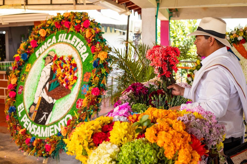 Hombre preparando arreglos florales coloridos, posiblemente para un festival o desfile. Incluye grandes coronas florales.