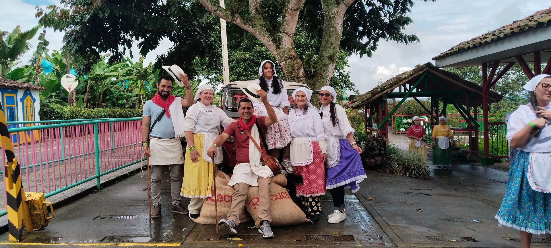 Un grupo de personas con trajes tradicionales posa con sacos. Se encuentran cerca de una valla colorida y árboles.