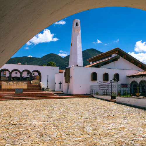 Un patio empedrado conduce a una iglesia blanca con un campanario alto y esbelto bajo un cielo azul brillante y nublado.