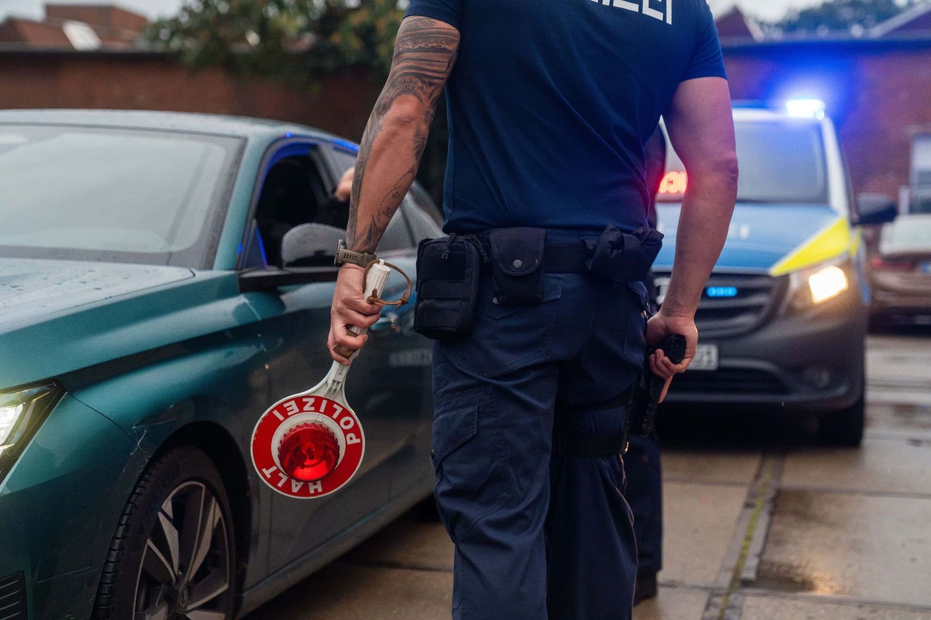 Polizist hält ein Stoppschild neben einem grünen Auto. Im Hintergrund sind blaue und rote Lichter zu sehen.