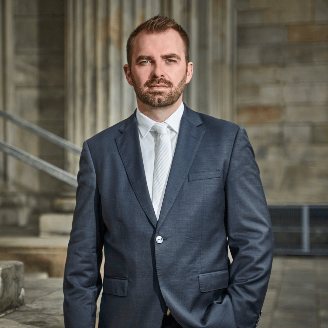 Mann im Anzug, Hände in den Taschen, steht vor einem Gebäude.