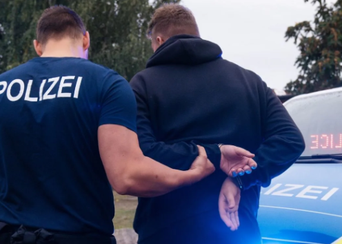Polizist nimmt eine Person fest, die Hände sind auf dem Rücken gefesselt, am Polizeifahrzeug blinken Blaulichter, im Freien.