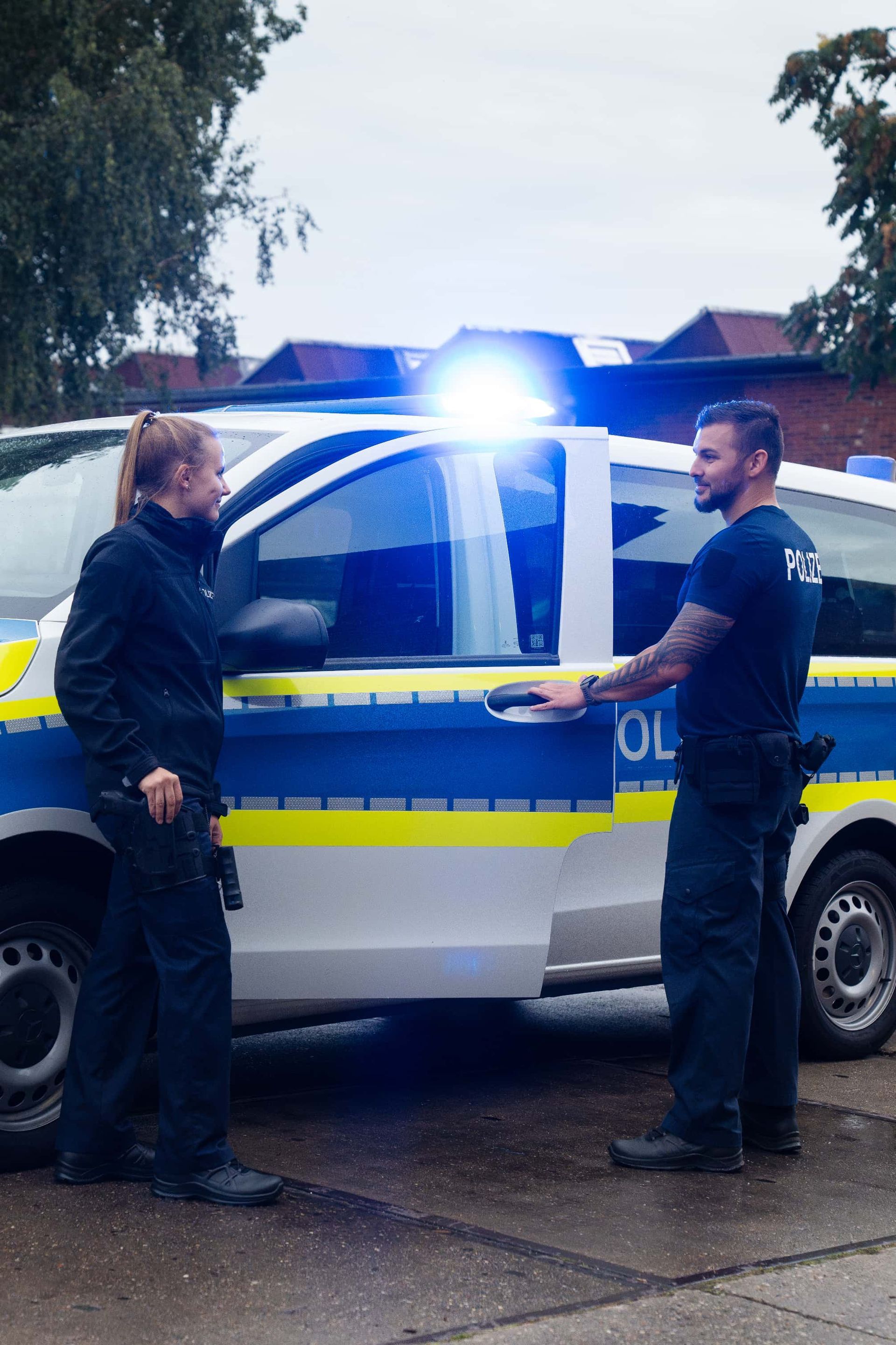 Zwei Polizisten stehen neben einem Polizeiwagen mit Blaulicht.