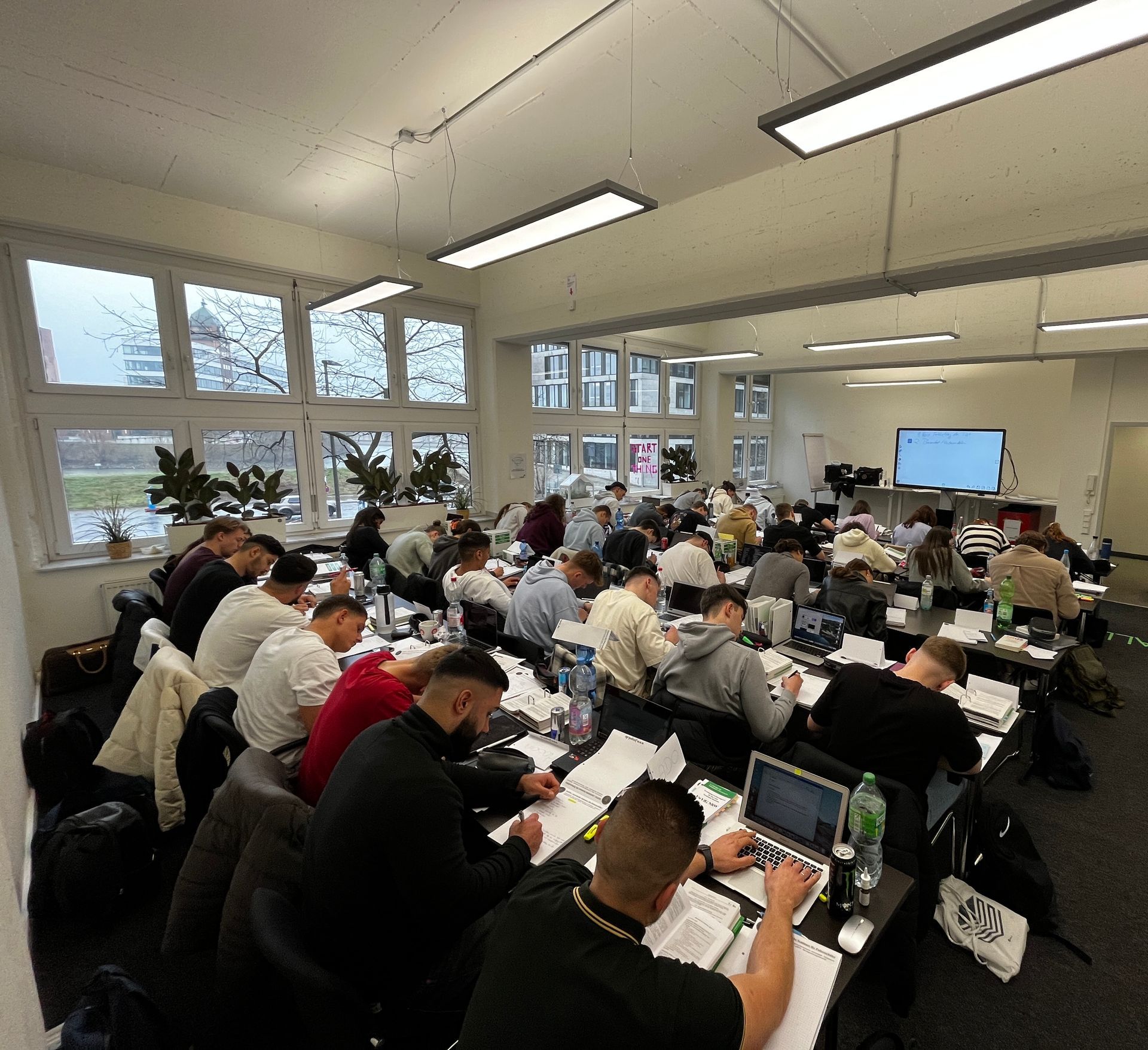 Großes Klassenzimmer mit vielen Leuten, die an Schreibtischen sitzen und an Papieren und Laptops arbeiten.