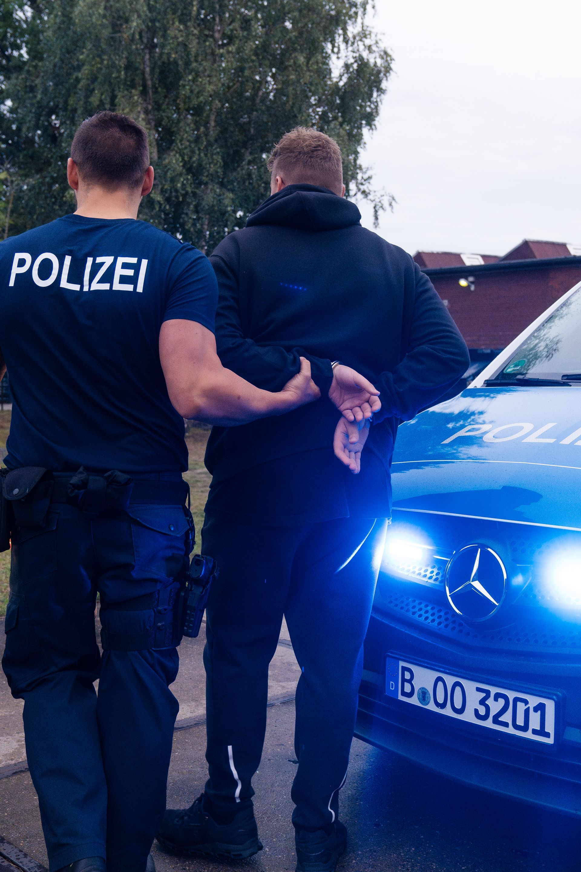 Ein Polizist nimmt einen Mann vor einem Polizeiauto fest. Der Polizist dreht ihm den Rücken zu.