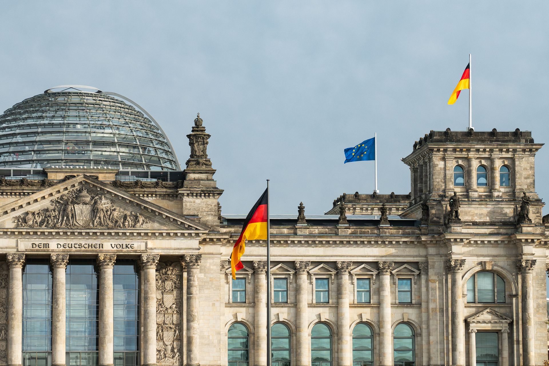 Reichstagsgebäude in Berlin mit wehenden deutschen und EU-Flaggen vor einem bewölkten Himmel.