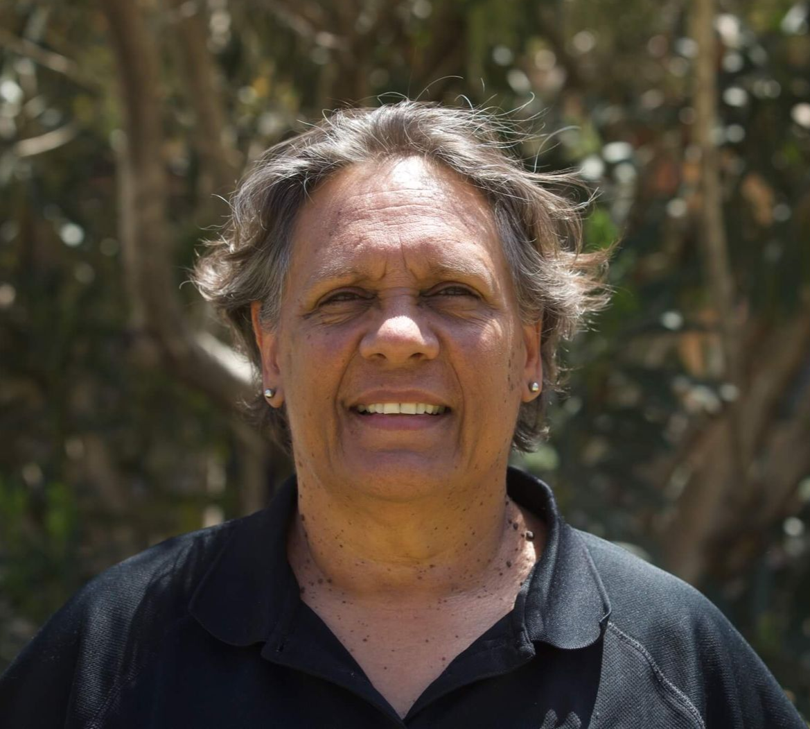 Person smiling, wearing a black shirt, against a blurred background of green foliage.