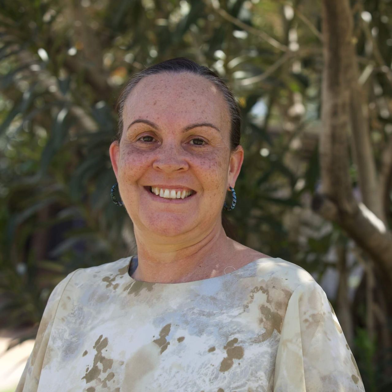 Woman smiles outdoors, wearing a light-colored patterned top, with trees in the background.
