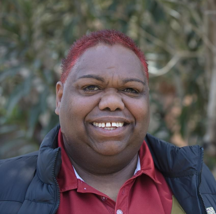 Person with red-dyed hair smiling, wearing a red collared shirt and black jacket, outdoors.
