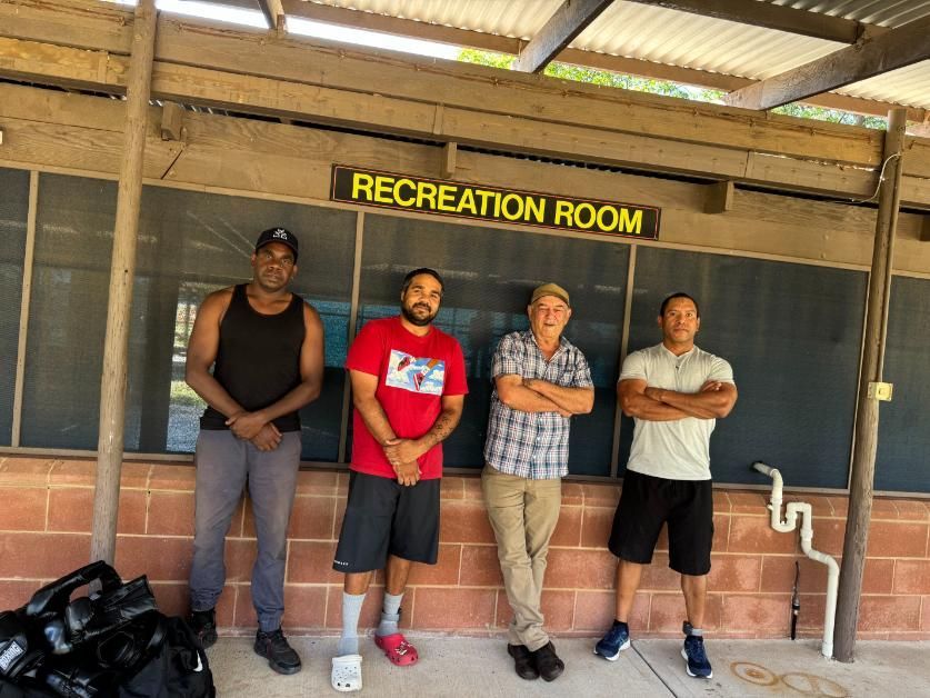 Four people stand in front of a "RECREATION ROOM" sign. Two men have crossed arms. Building with red brick.