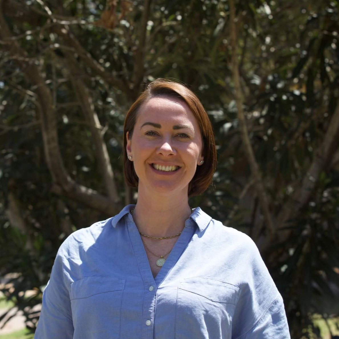 Woman with short brown hair smiles, wearing a light blue button-up shirt, outdoors in front of a tree.
