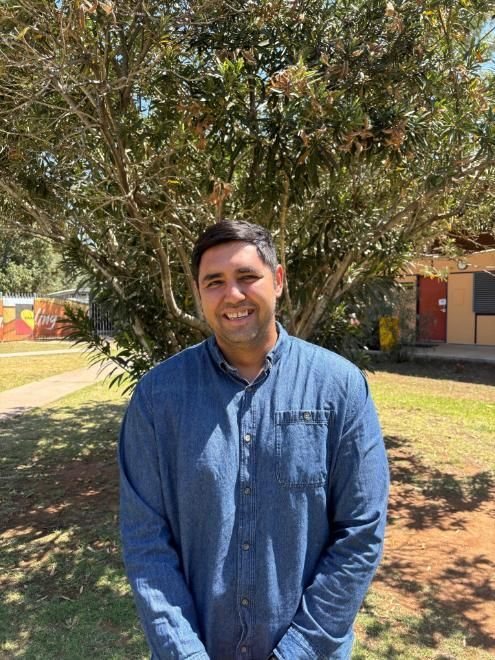 Man in blue shirt smiles in front of a tree and building.