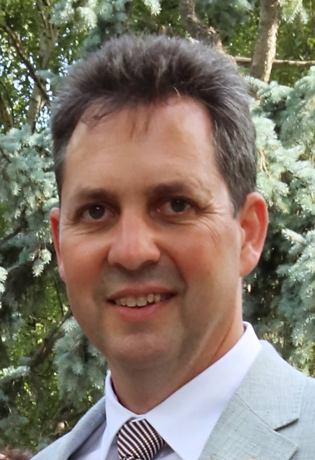Man in light suit smiling, standing outdoors near green foliage.