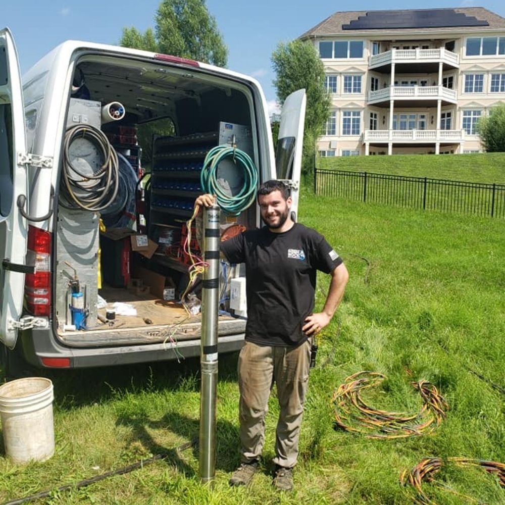 Man holding a pump, standing beside a van with equipment, on a grassy lawn, a house in the background.