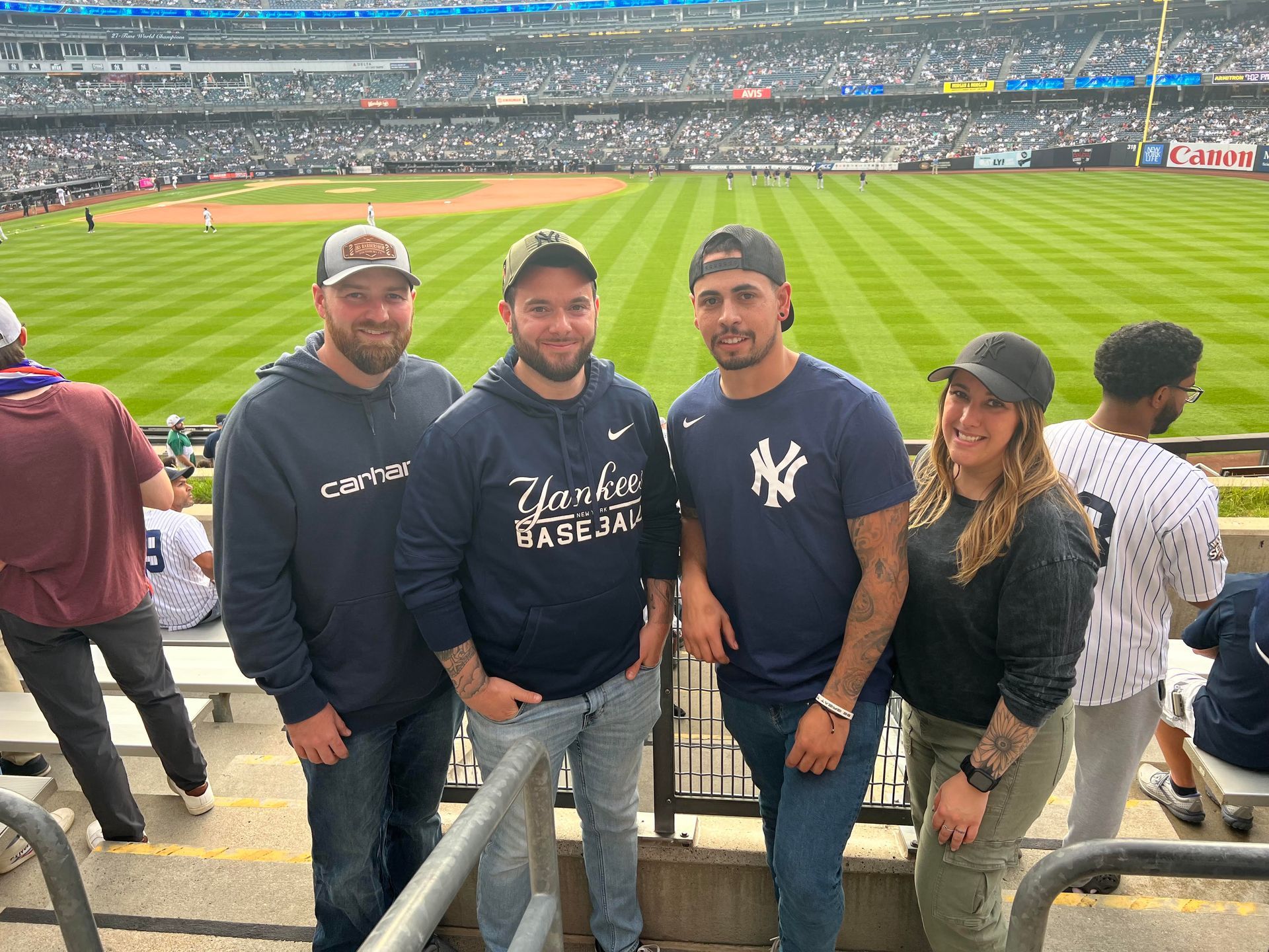 Four people at a Yankees baseball game, posing for a photo in the stands. Green field, blue and white colors.