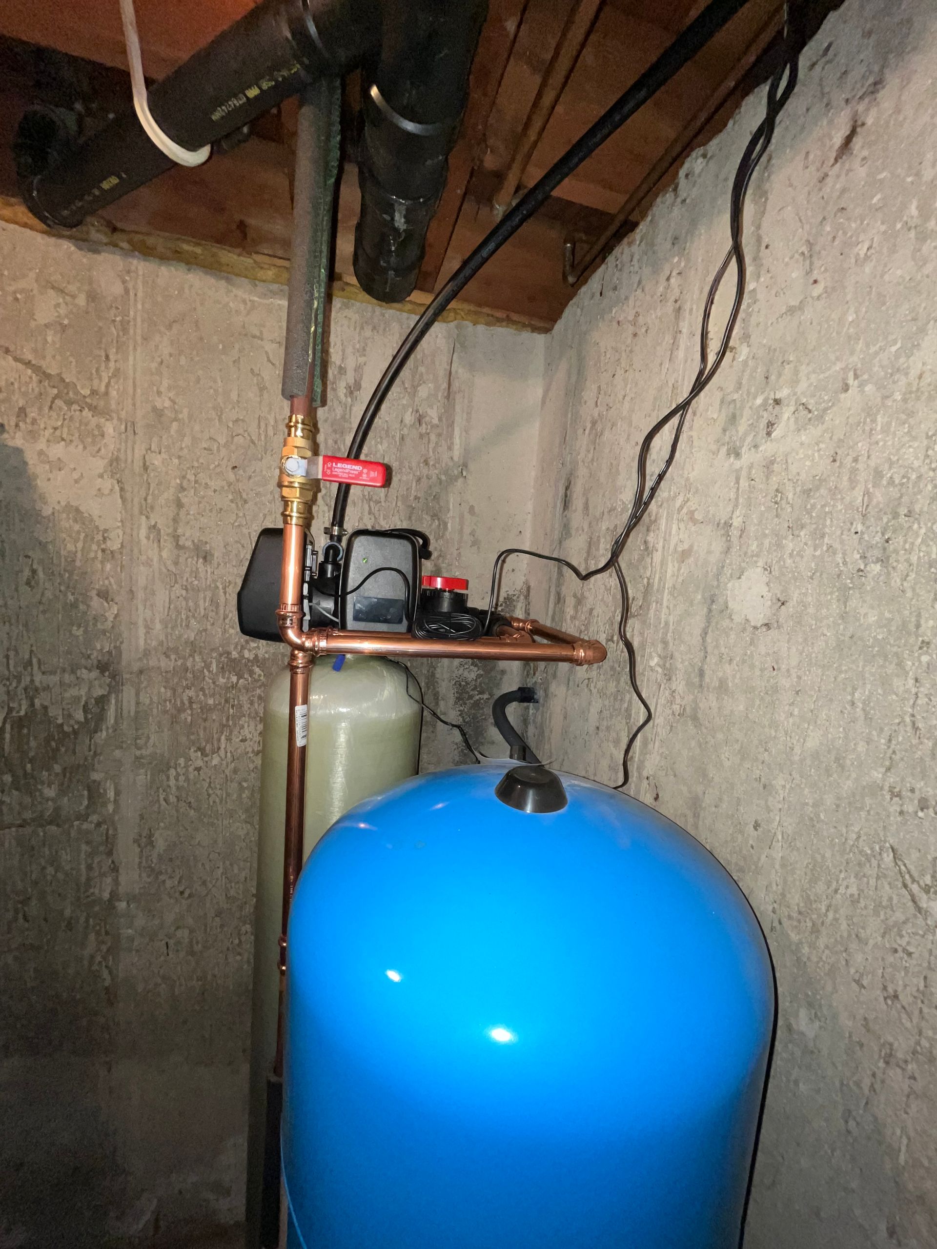 Blue water pressure tank, copper pipes, black pump, and filter unit in a basement setting.