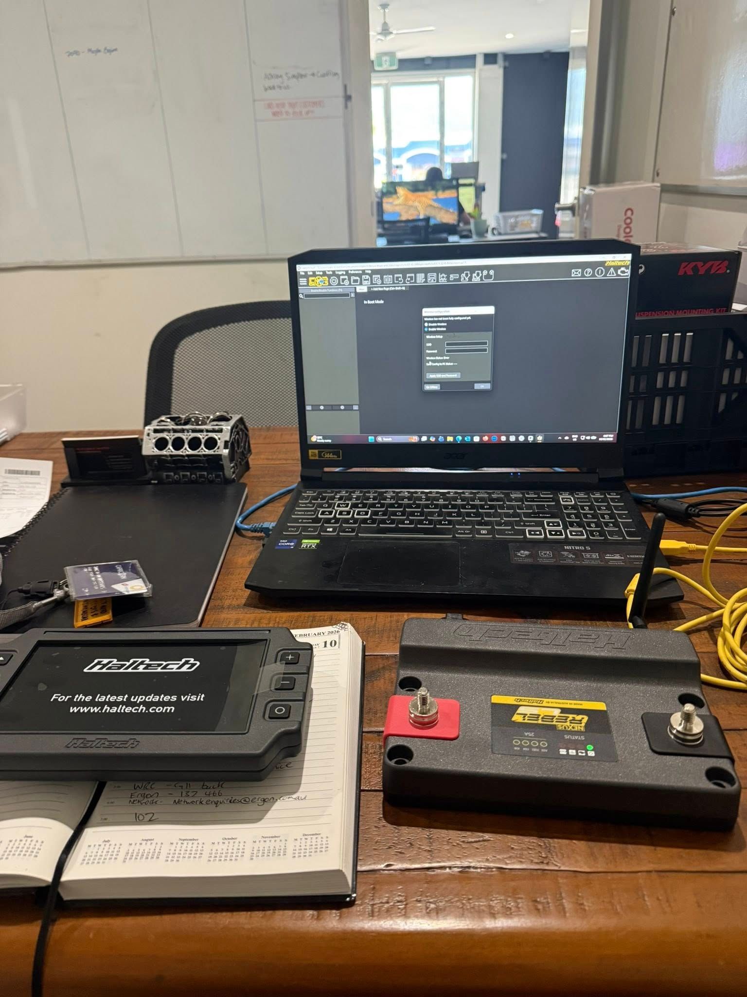 A laptop running software sits on a wooden desk next to a battery, a handheld device, and cables in an office workspace — Elite Fleet Auto in Cannonvale, QLD
